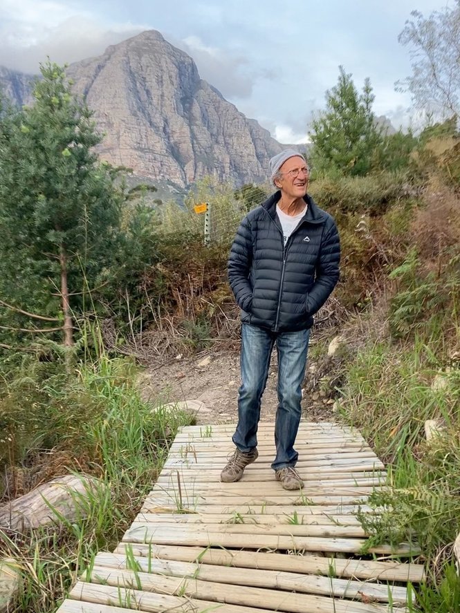 Dr Bernard Brom standing on a wooden boardwalk bridge, mountains behind